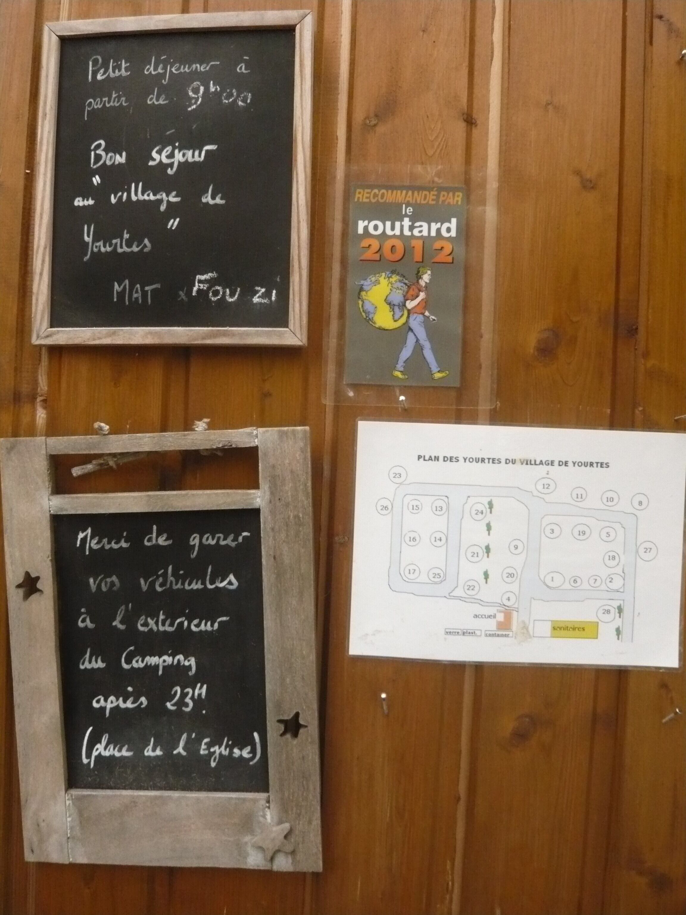 Eine Holzwand mit drei Elementen: einer Tafel mit einer Willkommensbotschaft, einem Buch mit dem Titel 'RECOMMANDÉ PAR le routard 2012' und einer Karte mit der Aufschrift 'PLAN DES YOURTES DU VILLAGE DE YOURTES'.