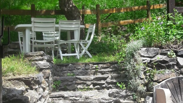 Una escala de pedra que condueix a una zona d'estar amb cadires blanques i una taula, envoltada de verdura i una tanca de fusta.
