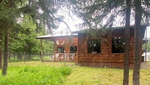 A rustic wooden cabin with a fenced patio area, surrounded by lush greenery and trees.