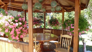 A wooden deck with a round table and chairs, surrounded by hanging flower baskets and a wooden pergola with hanging lanterns.