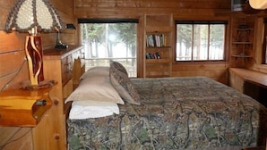 A wooden cabin bedroom with a bed, a lamp, a bookshelf, and a window with a view of trees.