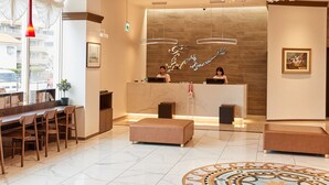Zona de recepción de hotel con suelo de mármol, sillas de madera, mostrador de recepción y una pared grande con un logotipo.