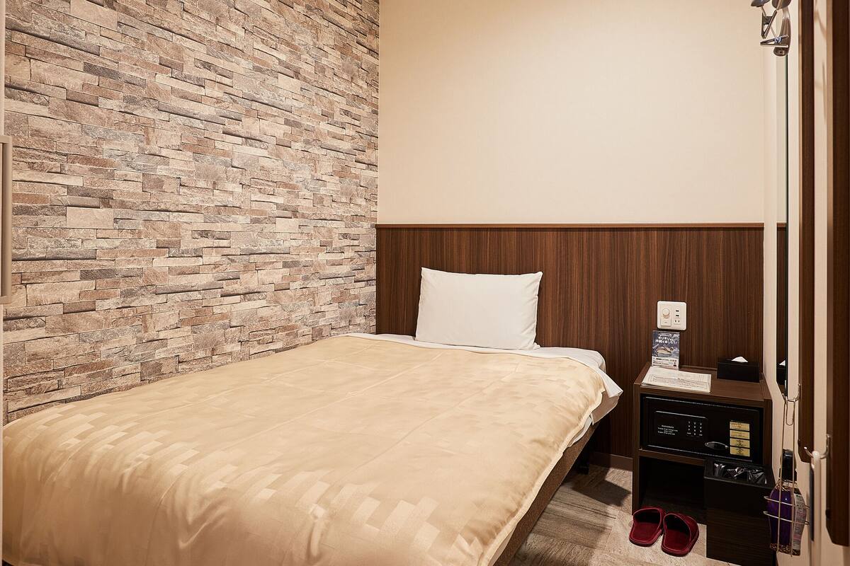 A hotel room with a stone-patterned wall, a wooden headboard, a bed with a beige bedspread, a nightstand with a lamp, and a pair of slippers on the floor.