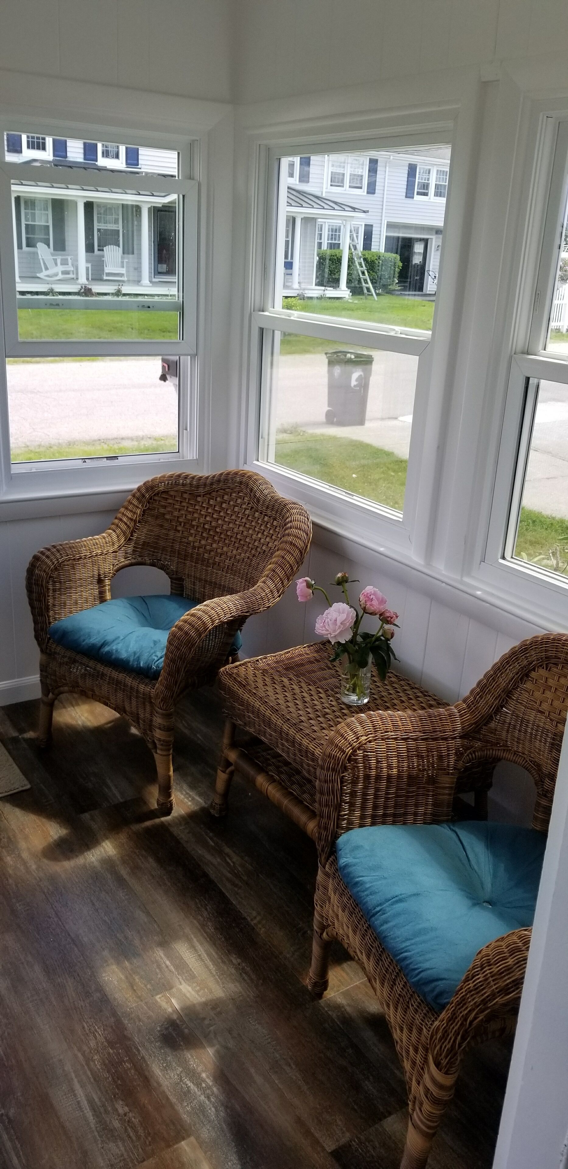 A room with two wicker chairs with blue cushions, a small table with a vase of flowers, and large windows offering a view of a residential area.