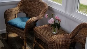 A room with two wicker chairs with blue cushions, a small table with a vase of flowers, and large windows offering a view of a residential area. - Beachside Family Getaway! 3 Doors From Beach Access. Fantastic!!!! (Biddeford)