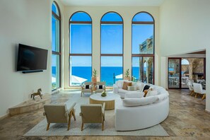 A spacious living room with a large white sectional sofa, a flat-screen TV mounted on the wall, and a view of the ocean through large arched windows. - Casa Rio de Luna: 7BR Ocean-View Villa with Pool, Rancho Cerro Colorado (Los Cabos)