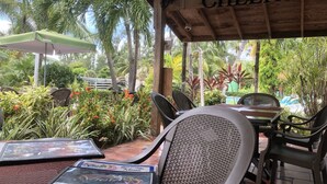 Un área de estar al aire libre con techo de madera y un letrero que dice 'CHEERS', rodeada de exuberante vegetación y una piscina al fondo.