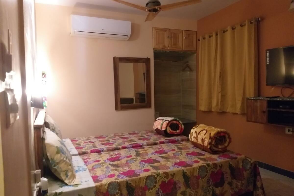 A bedroom with a bed, a ceiling fan, an air conditioner, a mirror, a television, and a window with curtains.