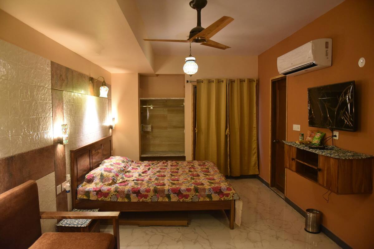 A bedroom with a bed, a ceiling fan, an air conditioner, a shower, and a small table with a lamp.