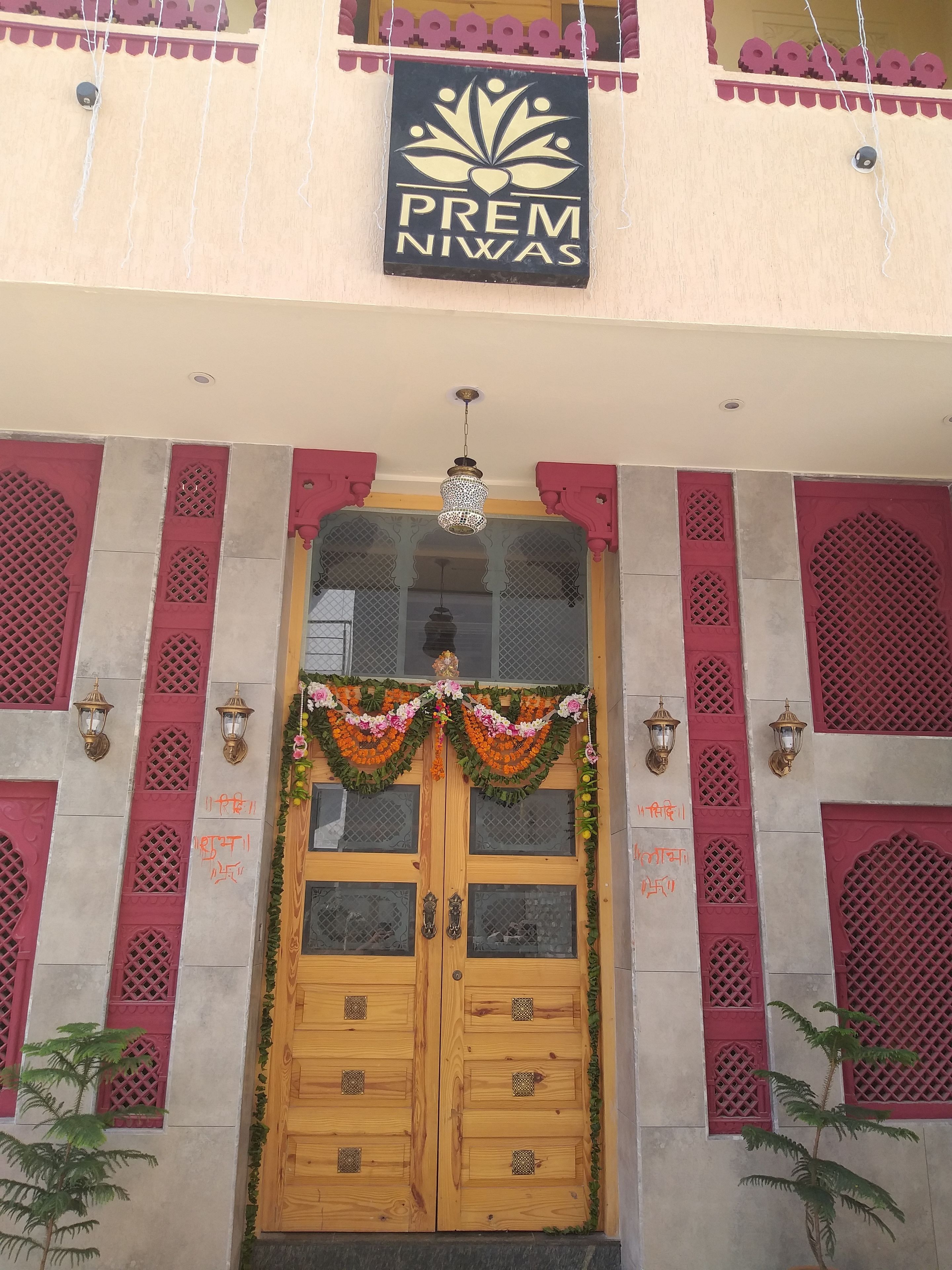 A hotel entrance with a wooden double door, a sign above reading "PREM NIWAS", and decorative garlands.