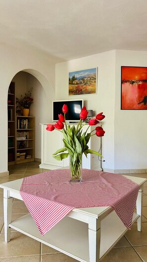 Un coin repas avec une table recouverte d’une nappe à rayures, un vase avec des fleurs rouges, une bibliothèque et un téléviseur en arrière-plan.