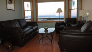 Ein Wohnzimmer mit braunem Ledersofa, einem Couchtisch und Blick auf ein Gewässer durch große Fenster.