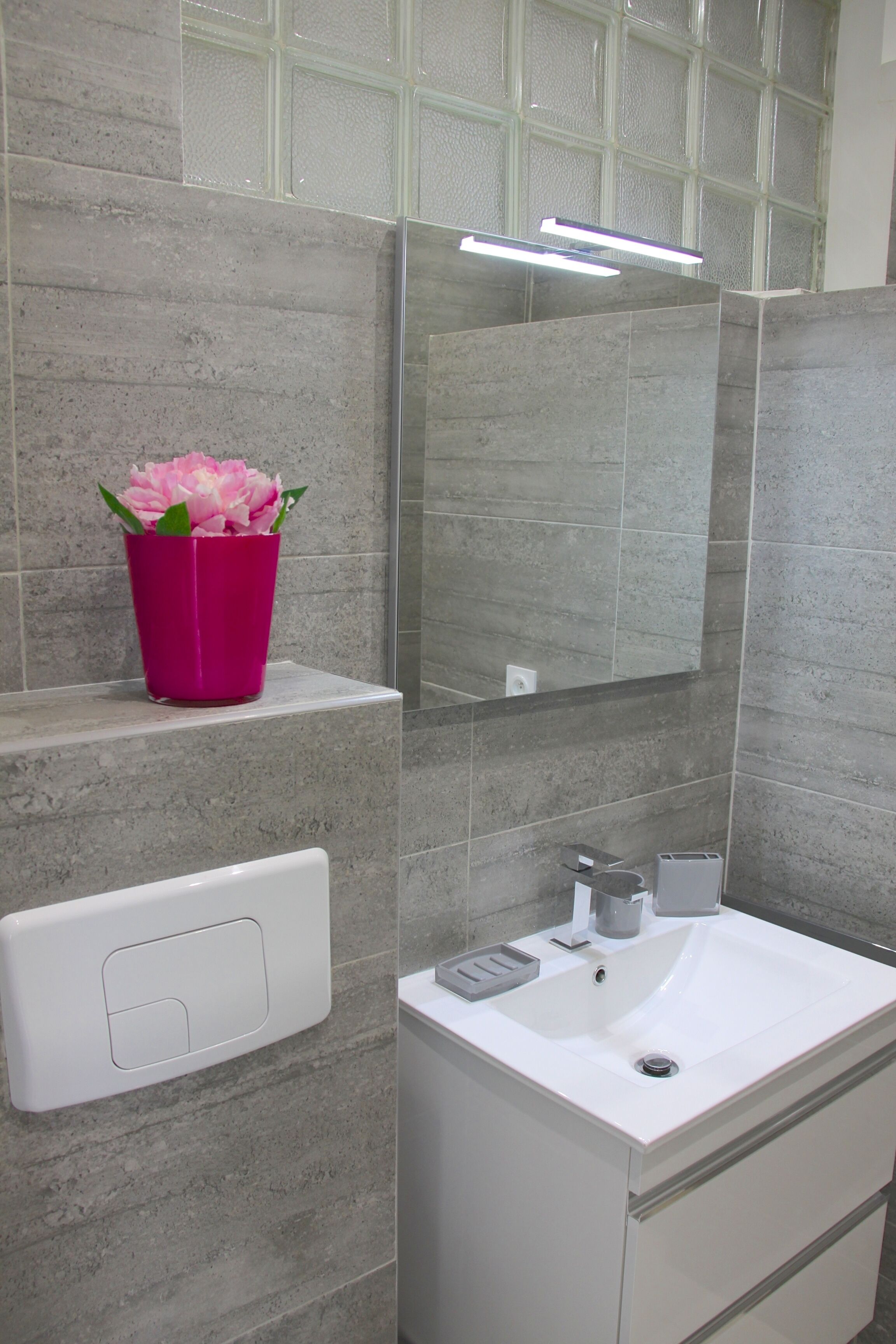 Une salle de bain avec un lavabo blanc, un vase avec une fleur rose et un miroir au-dessus du lavabo.