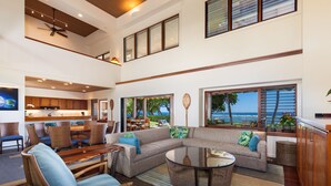 A spacious living area with a large sectional sofa, a round coffee table, and wicker chairs. There is a kitchen area with wooden cabinets and a dining table. Large windows offer views of the ocean.