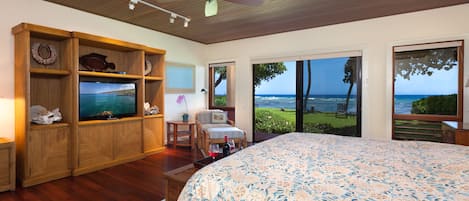 A bedroom with a large bed, wooden shelving, a television, and a view of the ocean through large sliding doors.