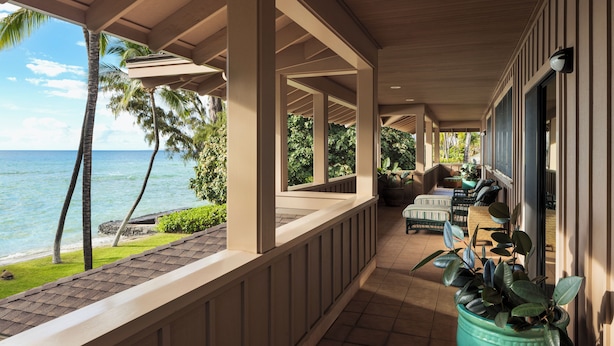A covered outdoor area with a view of the ocean, featuring a seating area with chairs and a table, surrounded by potted plants.