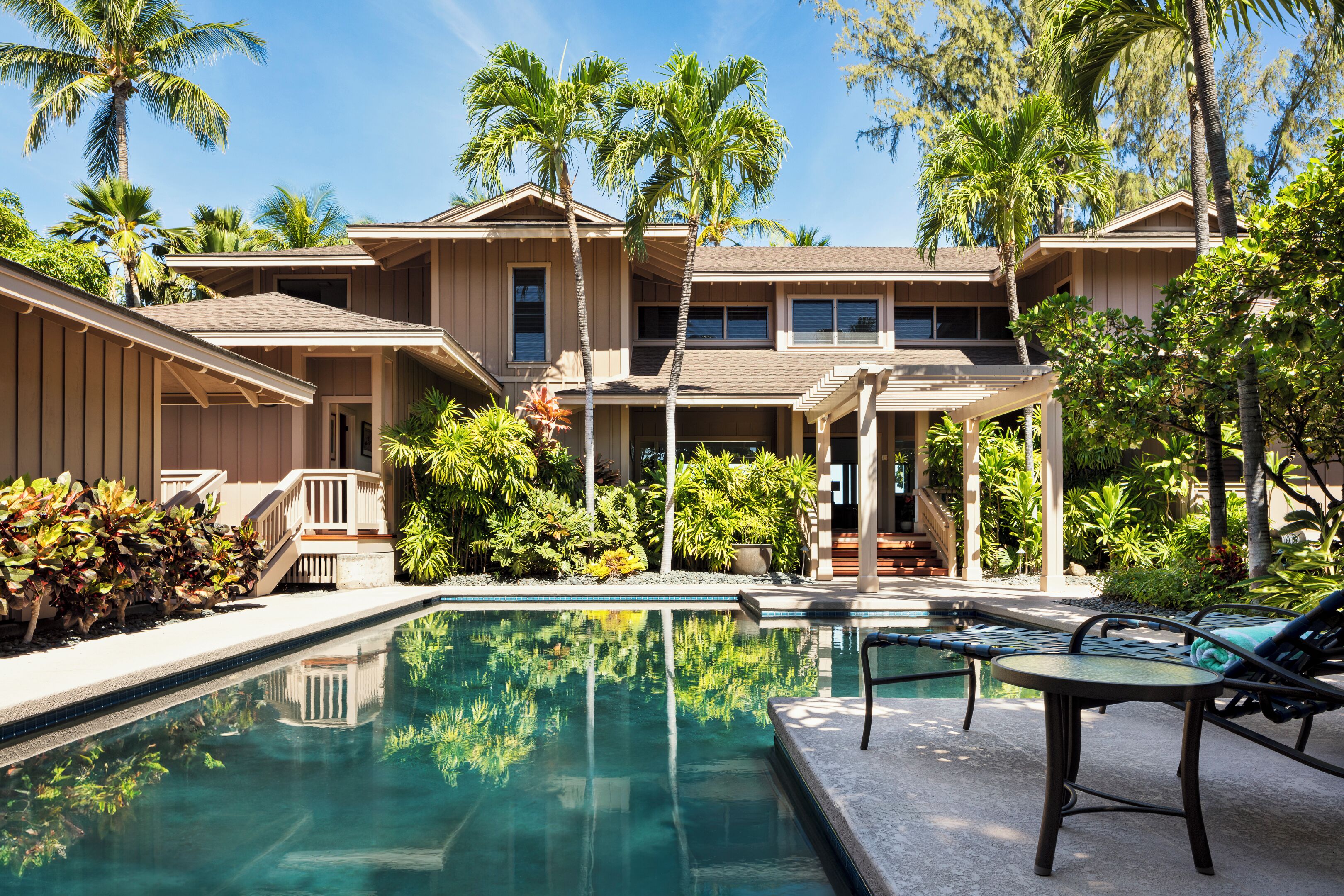 A two-story building with a swimming pool, surrounded by palm trees and various plants.
