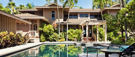 A two-story building with a swimming pool, surrounded by palm trees and various plants.