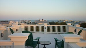 Een balkon met witte reling, groene stoelen en een tafeltje, met uitzicht op een kustgebied met gebouwen en de zee.