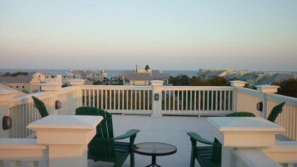A balcony with white railings, green chairs, and a small table, overlooking a coastal area with buildings and the sea. - Stunning Rooftop Views. Prime Island Location (Saint Simons Island)