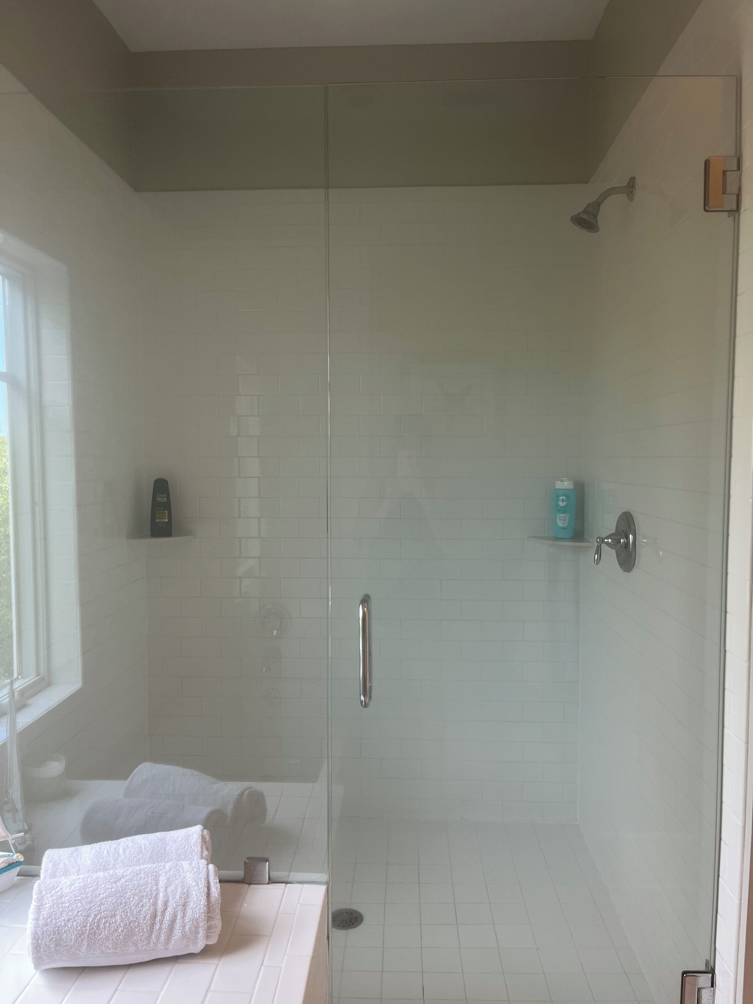A modern bathroom with a glass-enclosed shower, white tiled walls, and a window providing natural light.