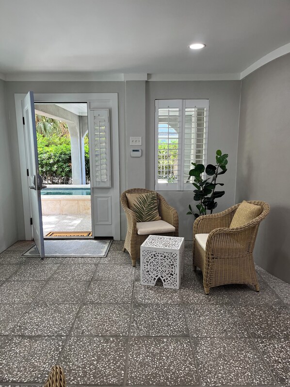 Een kamer met een betegelde vloer, twee rieten stoelen, een kleine witte tafel en een deur die naar buiten leidt.