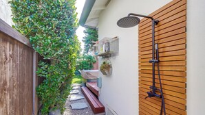 Un étroit sentier extérieur, agrémenté d’un banc en bois, d’une douche murale en bois et d’un petit espace jardin avec des plantes et une cage à oiseaux.