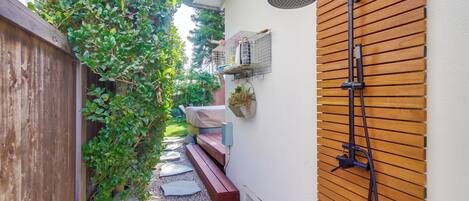 Un sendero estrecho al aire libre con un banco de madera, una ducha de madera montada en la pared y un pequeño jardín con plantas y una jaula de pájaros.