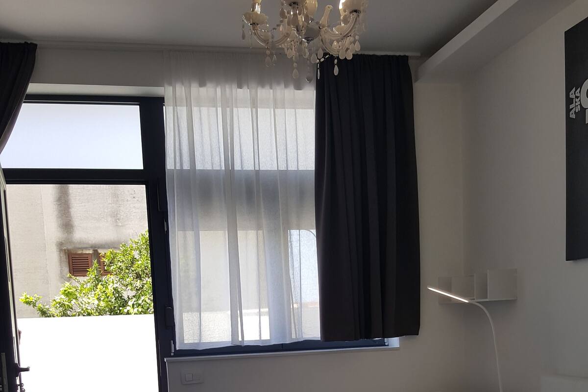 Habitación de hotel moderna con una cama grande, una mesita de noche, un escritorio, una silla, una araña y una ventana con cortinas.