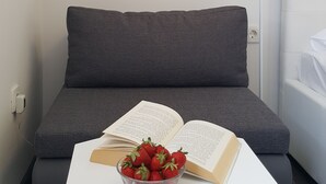 Quarto moderno com sofá cinzento, uma mesinha branca com uma tigela de morangos e um livro aberto, uma luminária de cabeceira e uma prateleira branca acima do sofá.