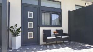 Fachada moderna com porta cinza escura, janela com cortinas e área de estar com duas cadeiras e uma pequena mesa.