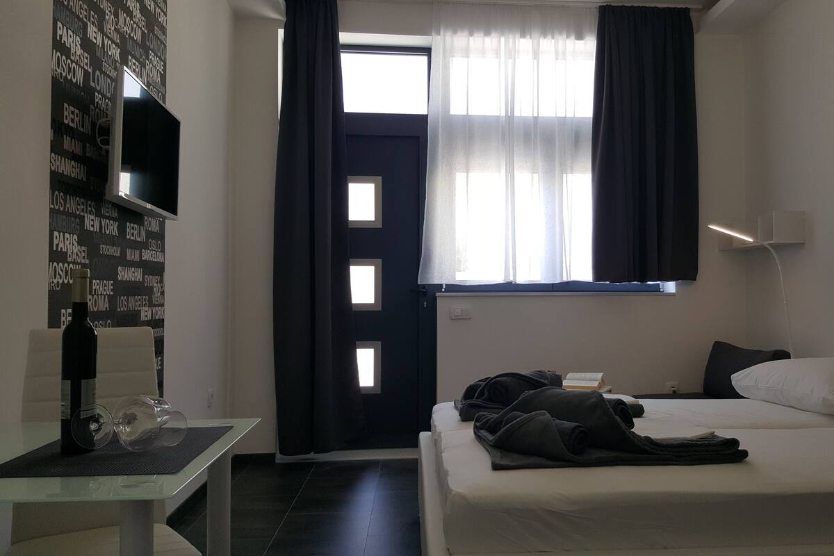 Habitación de hotel con cama, una mesa con botella y vasos, una ventana con cortinas y una pared con nombres de ciudades.
