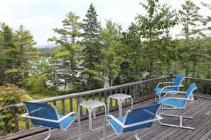 A deck with blue chairs and a small table, overlooking a forested area and a body of water. - Beautiful Seaside Home with Sweeping Views - Weekly Discounts Available! (Mt Desert)