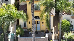 A yellow building with a blue door, surrounded by palm trees and potted plants. - Come stay at our spacious beach house! (Oceanside)