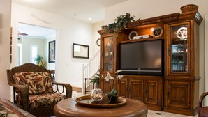 A living room with a large flat-screen TV, a floral armchair, a leather ottoman, and a wooden entertainment center. - Come stay at our spacious beach house! (Oceanside)