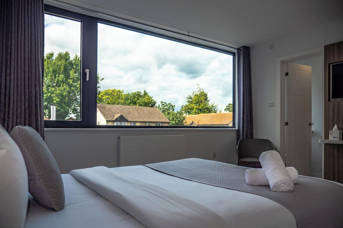 A hotel room with a large window, a bed with white linens, a chair, and a TV on the wall.