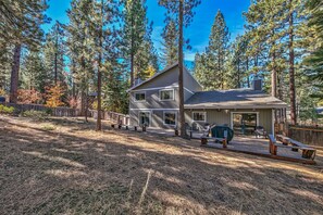 A two-story house with a deck, surrounded by tall pine trees.