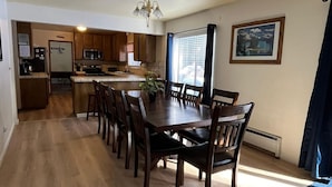 A spacious dining area with a long wooden table and chairs, a kitchen in the background, and a large window with curtains. - Meadow Drive Home In Beautiful Lake Tahoe Nevada (Stateline)