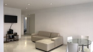 En moderne stue med en beige hjørnesofa, et spisebord med glasplade og hvide stole, samt et fladskærms-tv monteret på væggen.