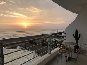 Een balkon met uitzicht op de oceaan bij zonsondergang, een cactusplant en een kleine tafel met stoelen.