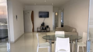 En moderne stue med et glasbord, hvide stole, en beige sofa, et fladskærms-tv monteret på væggen og en trædør med nummer 301.