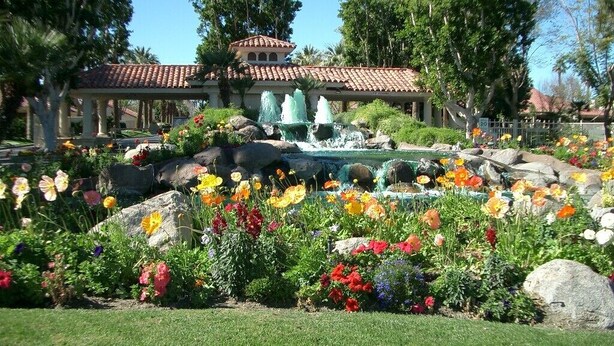 A garden with a fountain, colorful flowers, and a building with a tiled roof. - Green Vistas on the Tenth Tee is a one story end unit with wall to wall windows  (Palm Desert)