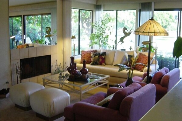 A modern living room with a sofa, ottoman, and coffee table. There is a fireplace, large windows, and a lamp.