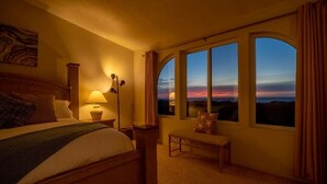 A hotel room with a large bed, a bedside table with a lamp, a bench, and a view of the sunset through arched windows.