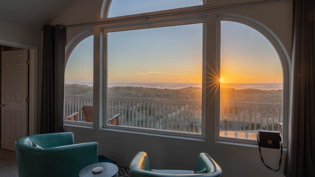 A room with a large arched window overlooking a beach at sunset, featuring two green chairs and a small round table.
