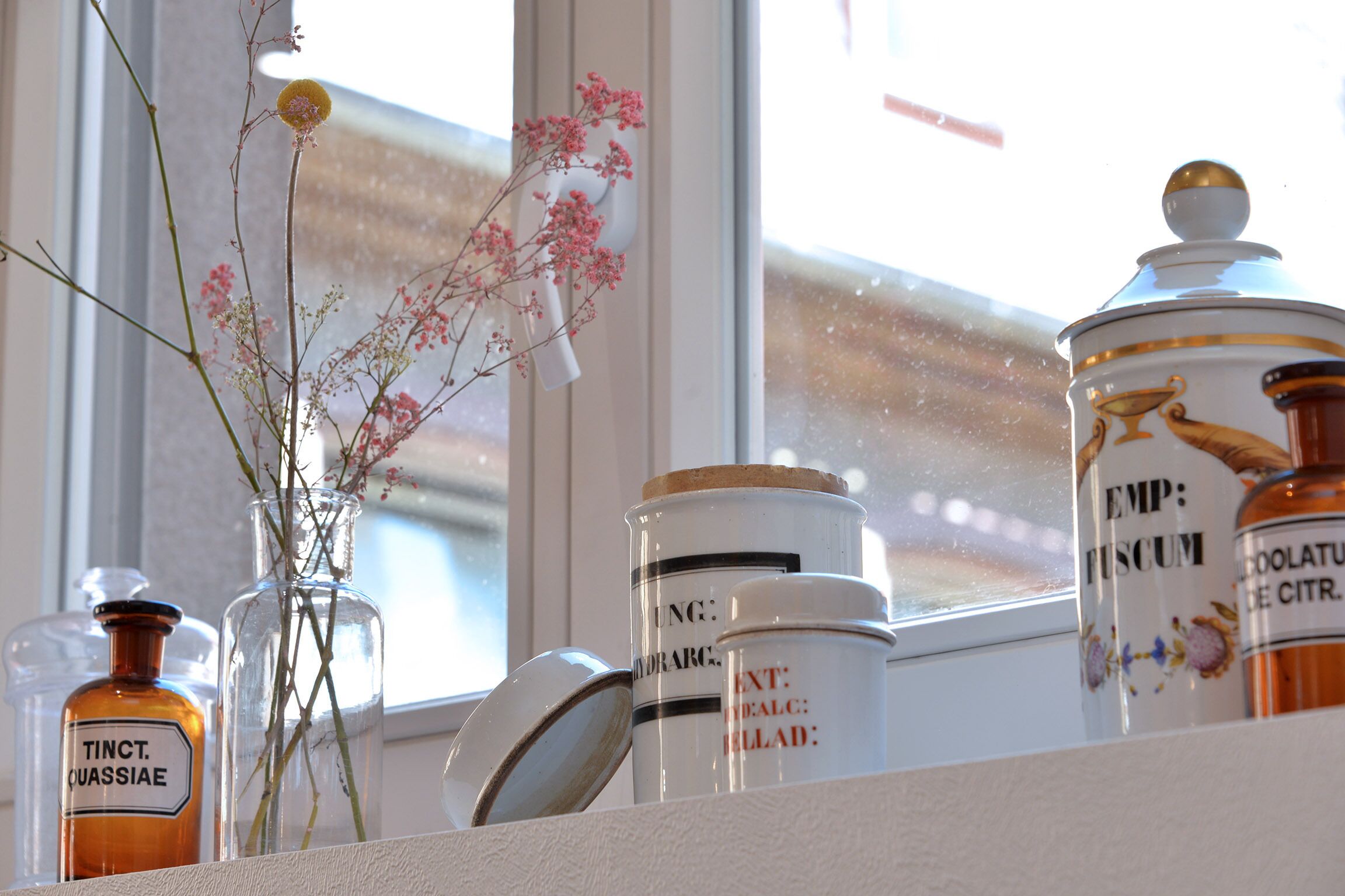 Uno scaffale con diversi contenitori di vetro, alcuni etichettati, e un vaso con fiori rosa.