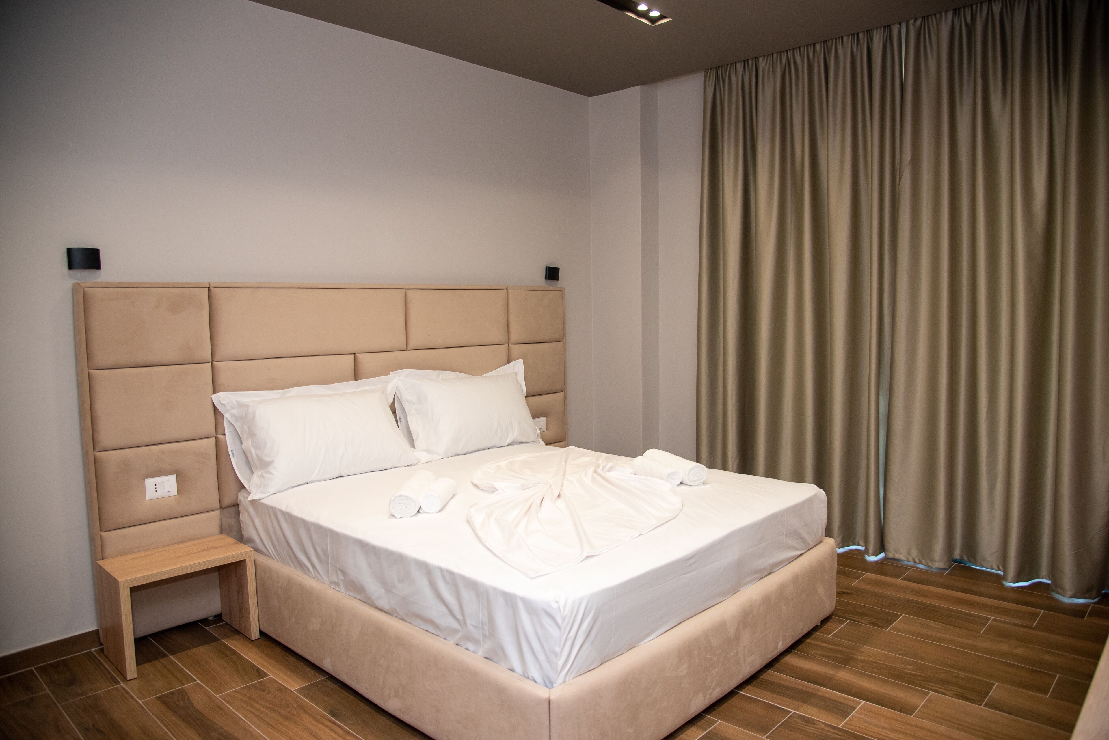 A neatly made bed with white linens and pillows, a wooden nightstand, and a large window with curtains.