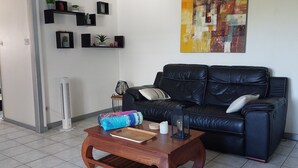 A living room with a black leather sofa, a wooden coffee table, a standing fan, and a wall-mounted shelf with decorative items. - Beautiful T2 apartment Clear view between sea and mountain near the lagoon (La Saline-Les-Bains)