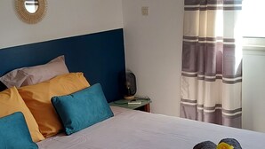 A neatly made bed with a light-colored bedspread and pillows in blue and yellow. A decorative mirror hangs on the wall, and a ceiling fan is visible. - Beautiful T2 apartment Clear view between sea and mountain near the lagoon (La Saline-Les-Bains)