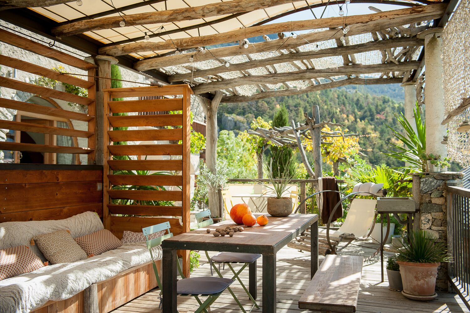 A wooden deck with a sofa and table, surrounded by potted plants and a view of a mountainous landscape.